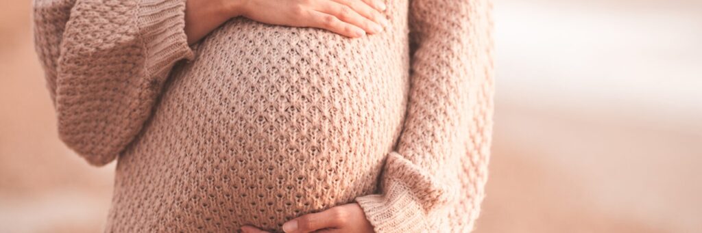 Pregnant woman wearing cozy knitted sweater holding tummy with hands outdoors close up. Motherhood. Maternity. Heathy lifestyle.