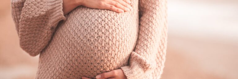 Pregnant woman wearing cozy knitted sweater holding tummy with hands outdoors close up. Motherhood. Maternity. Heathy lifestyle.